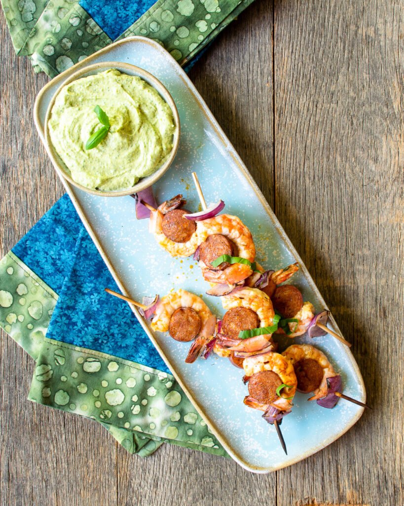 Grilled shrimp chorizo skewers on a blue rectangular plate with a dip and blue and green napkins.