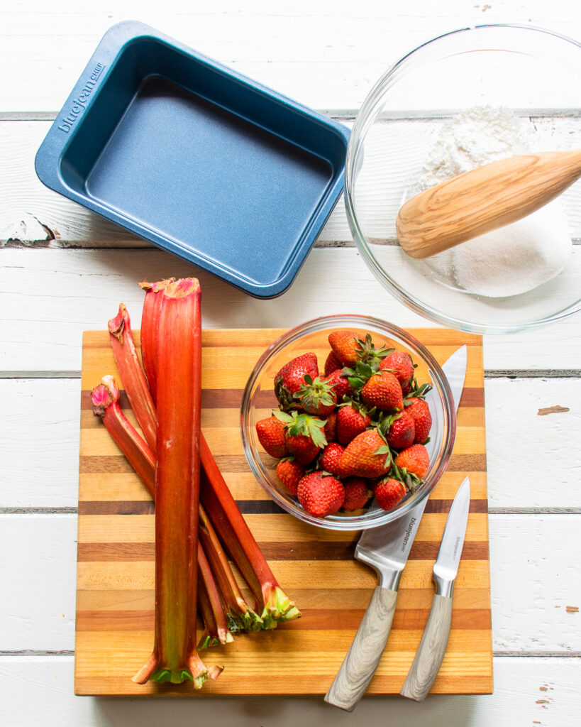 Rhubarb and strawberries on a wooden board with knives, bowls and a pan nearby. 