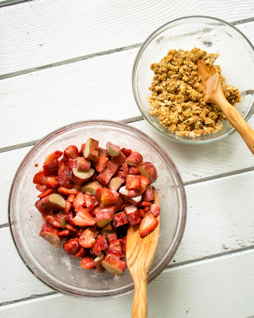 Strawberries and rhubarb in a bowl with a wooden spoon and a second bowl with crumble topping on a white wooden table.