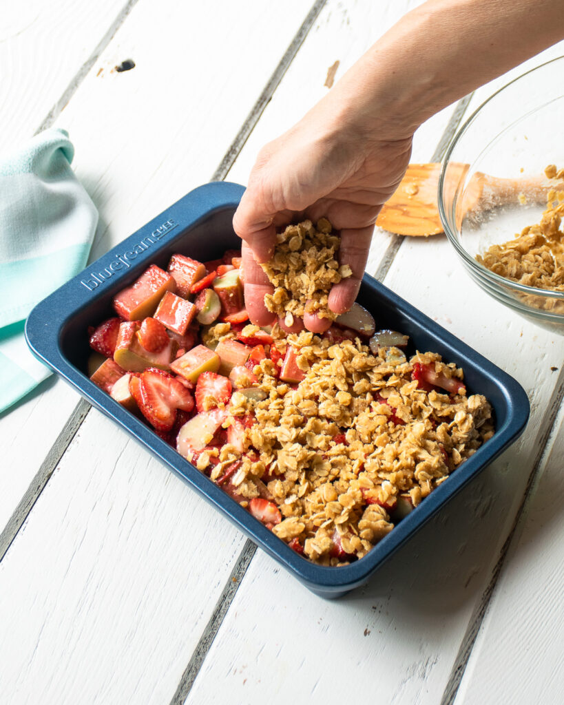 A hand spreading crumble topping on a strawberry rhubarb crisp in a blue baking pan.