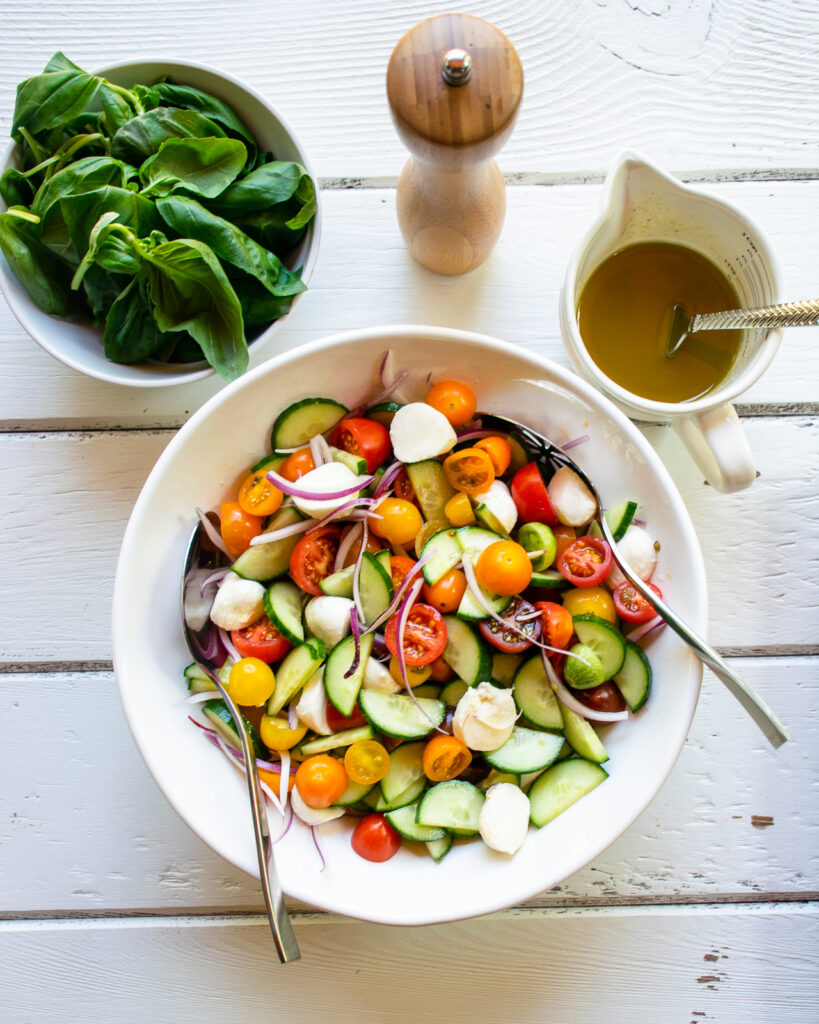 Tomato cucumber salad in a white bowl with dressing, basil and a pepper grinder near by.