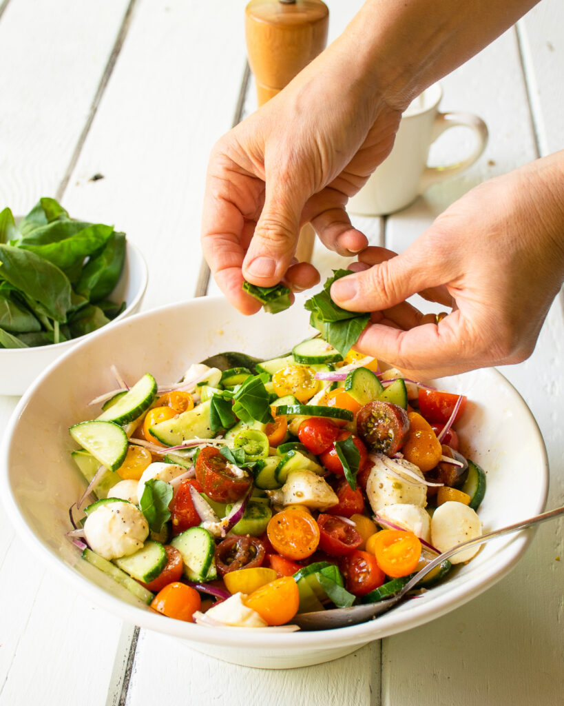Hands tearing basil over a tomato cucumber salad.