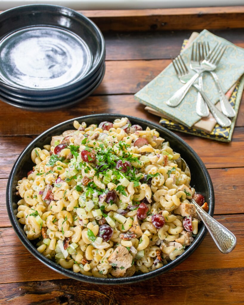 Chicken macaroni salad in a wide bowl on a wooden table with a stack of shallow bowls, napkins and forks.