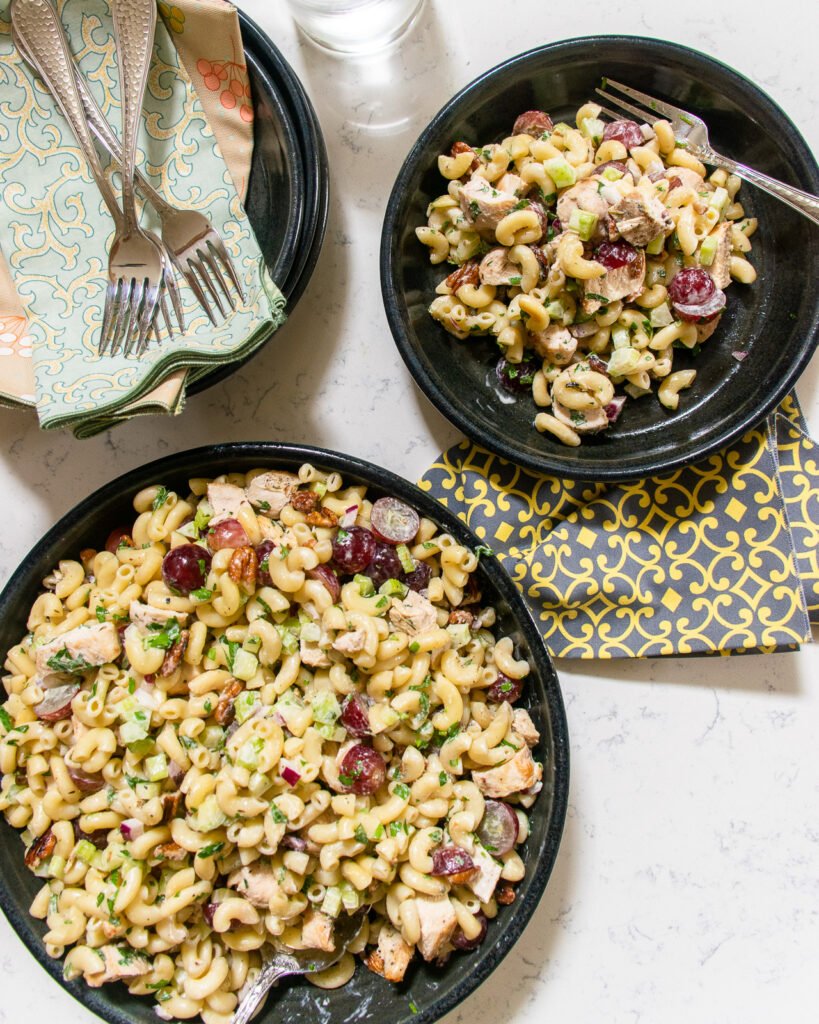 A serving of chicken macaroni salad next to a larger bowl with napkins and forks on the countertop.