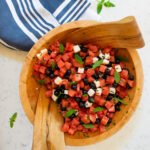Watermelon feta salad with blueberries in a wooden salad bowl with a blue and white striped towel next to it.