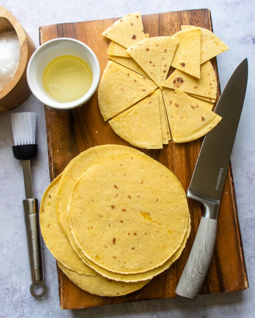 Corn tortillas on a wooden cutting board, some cut into wedges with a chef's knife, a bowl of oil and a basting brush.