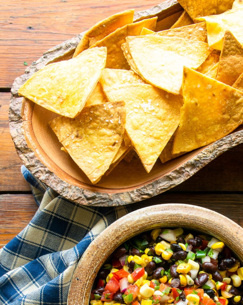 Tortilla chips in a wooden bowl on a wooden table with a bowl of salsa and a blue checkered napkin.