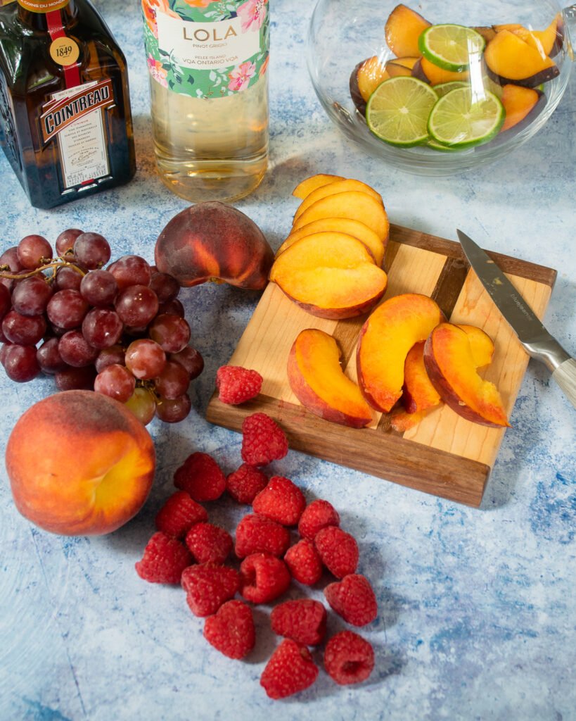 PEaches cut up on a little wooden cutting board with raspberries and grapes next to it and a pitcher with lime slices in the background.