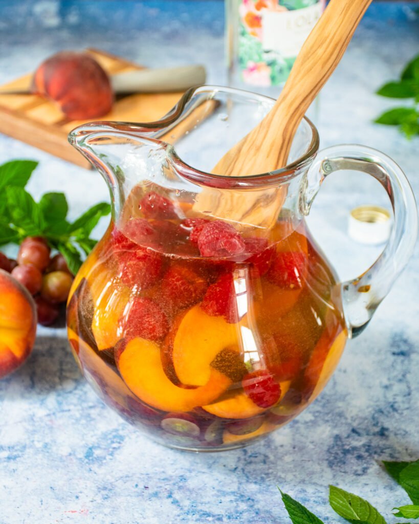 A pitcher of white wine sangria in a glass pitcher with a wooden spoon sticking out of it.