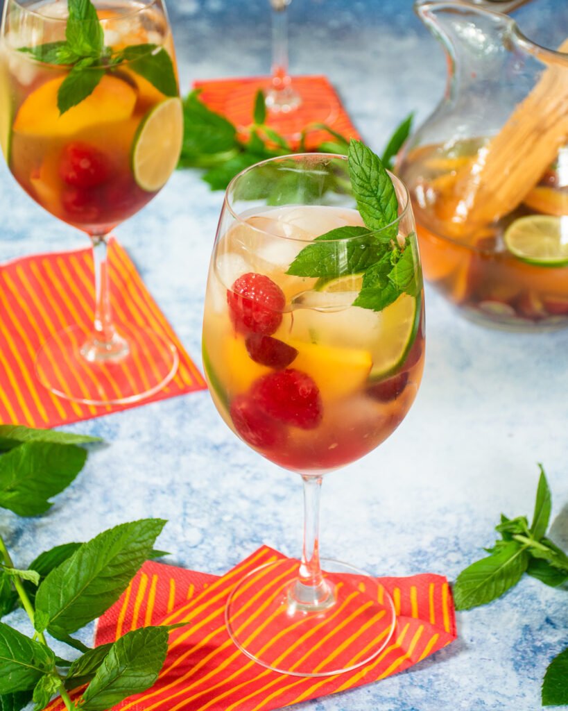 A glass of white wine sangria on a red and yellow napkin with a pitcher and another wine glass in the background.