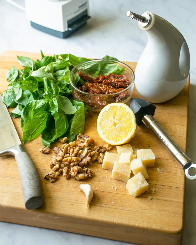 Ingredients on a cutting board - parmesan cheese, walnuts, sun-dried tomatoes, lemon, basil and a cruet of olive oil