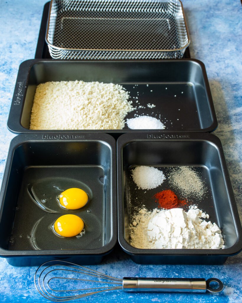 Baking pans on a blue counter with dredging ingredients - a pan of beaten egg, a pan of seasoned flour and a larger pan of breadcrumbs.