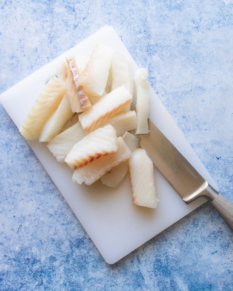 Cod pieces cut up on a white cutting board with a knife next to them.