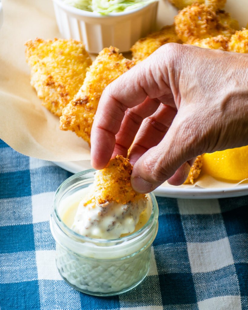 A hand dipping a fish stick into a jar of tartar sauce with a plate of fish sticks in the background.