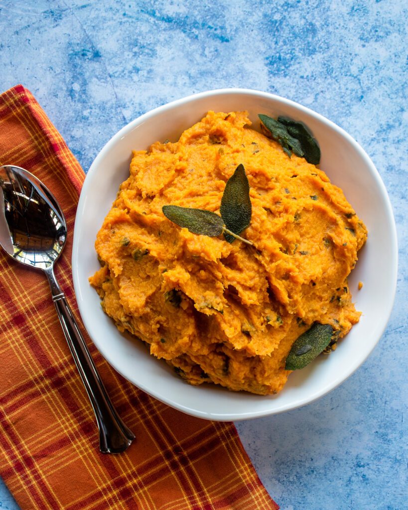 Looking down into a white bowl with mashed sweet potatoes decorated with fried sage leaves, a soon and an orange plaid kitchen towel.