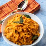 A white bowl with mashed sweet potatoes decorated with fried sage leaves, a soon and an orange plaid kitchen towel.