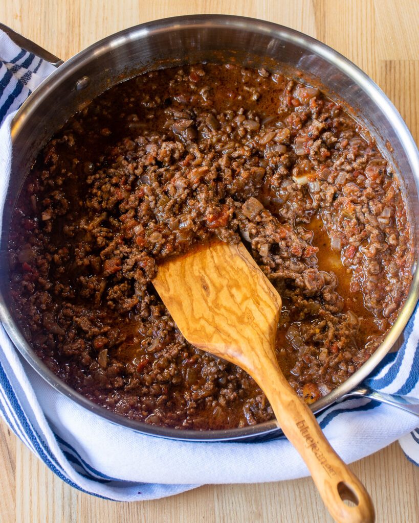 Meat sauce in a stainless steel saute pan with a wooden spoon and a blue and white striped towel.
