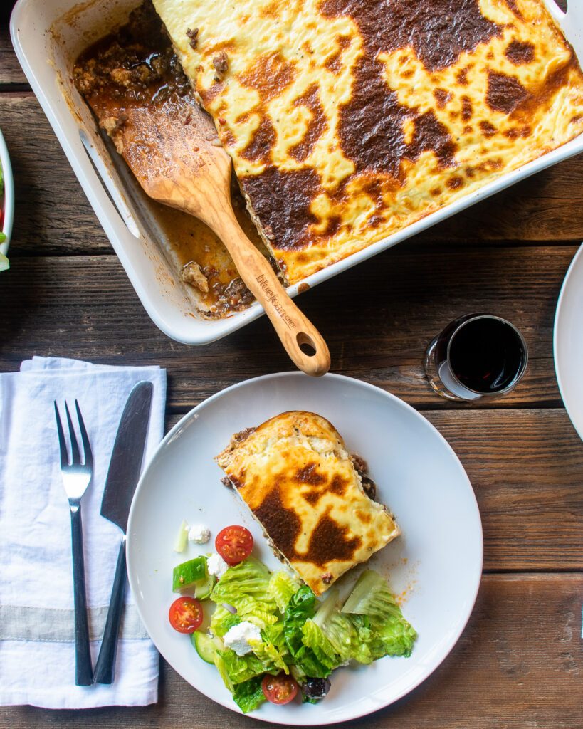 Looking down on a dinner table with a white casserole pan of moussaka, a white plate of greek salad and a white dinner plate of a portion of moussaka with salad.