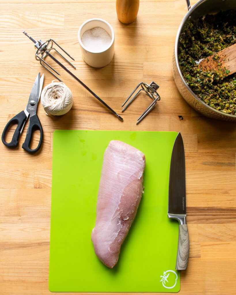 Turkey breast on a green cutting board with a chef's knife, rotisserie spit, scissors and kitchen twine near by.