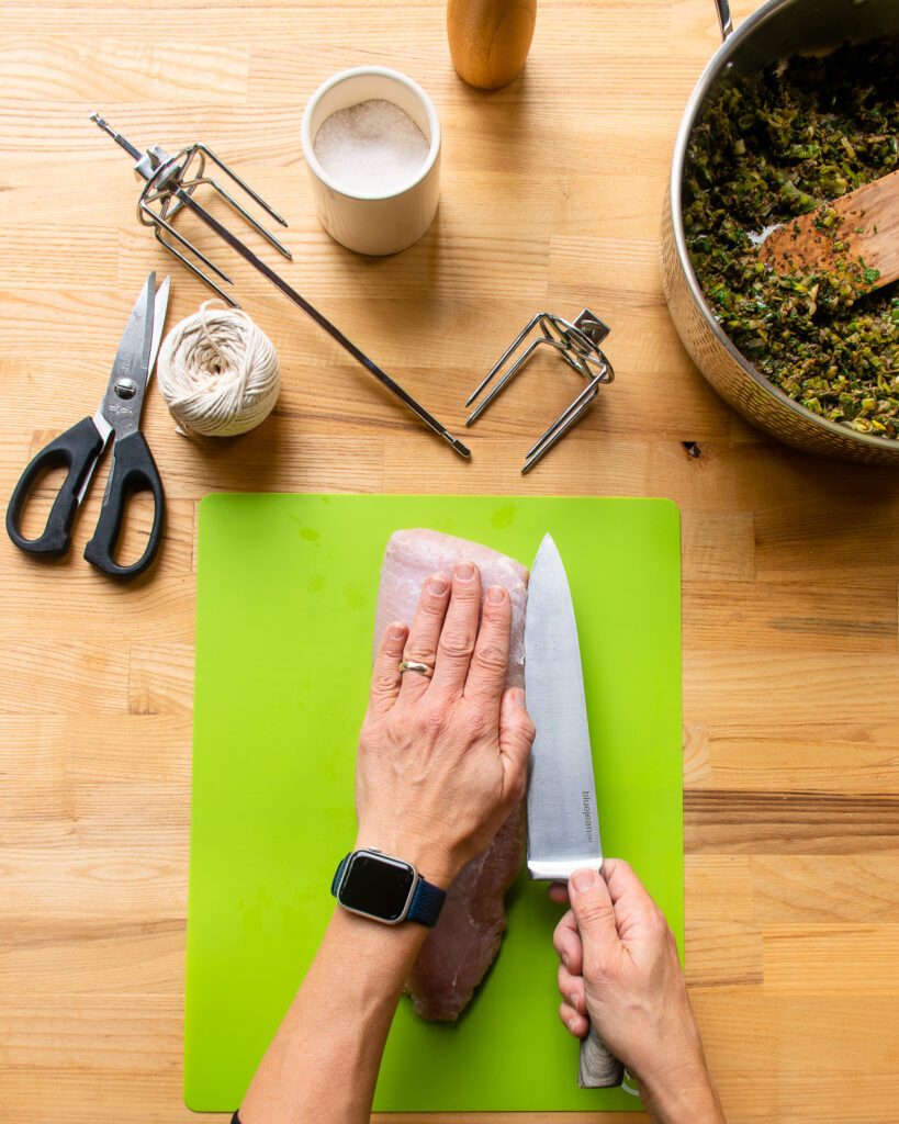 A hand pressing down on a turkey breast while a knife slices into the breast parallel to the cutting board.