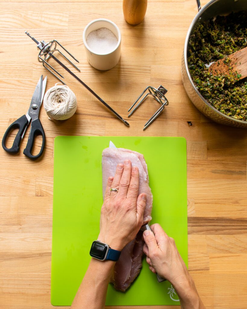 A hand pressing down on a turkey breast while a knife slices into the breast parallel to the cutting board.
