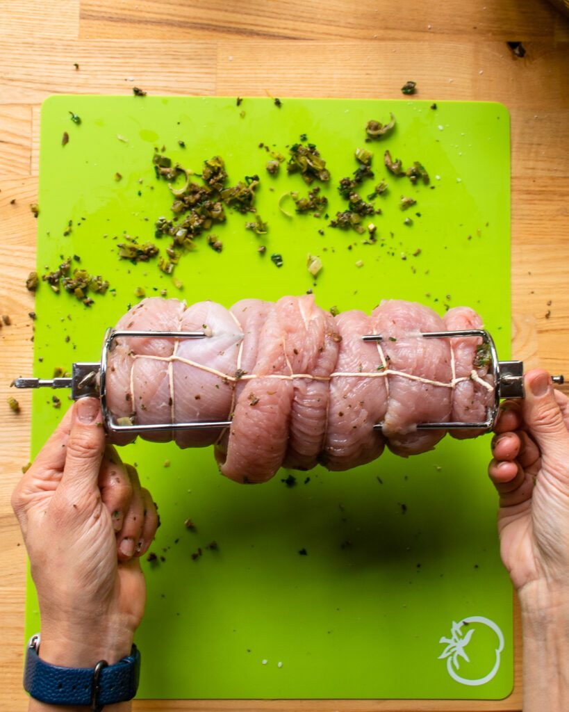 Two hands holding the ends of a rotisserie spit with a turkey breast roulade on it. 