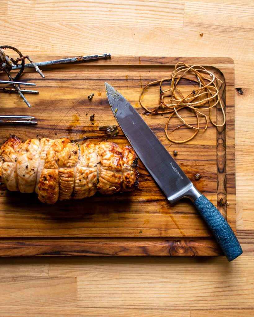 roasted turkey roulade on a cutting board with a chef's knife, the rotisserie spit and prongs and some discarded kitchen twine.