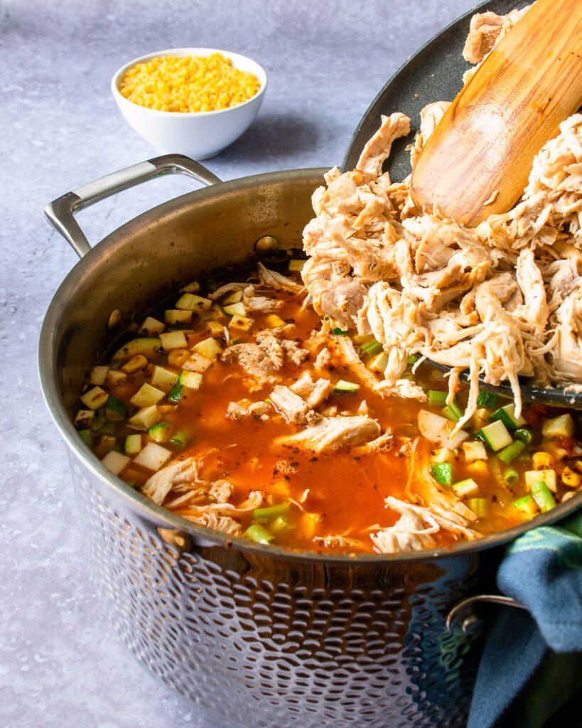 A wooden spoon pushing shredded chicken off a plate and into a hammered stainless steel stockpot with vegetable soup.