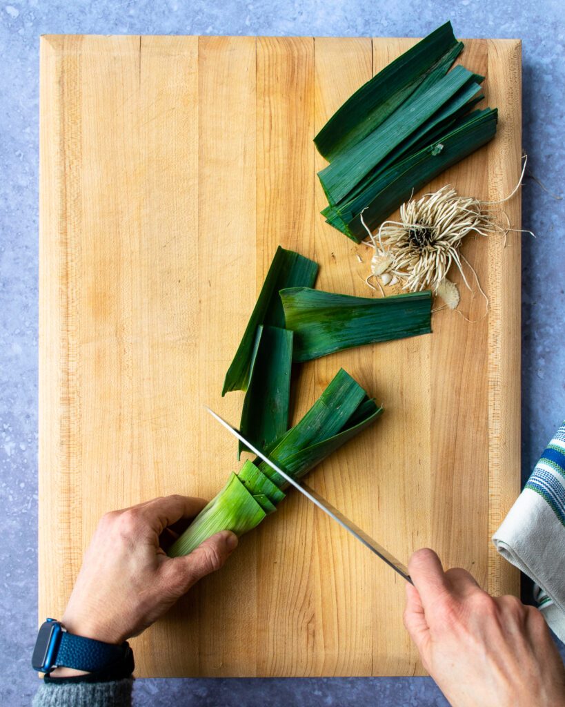 Hands cutting the dark green leaves off a leek.