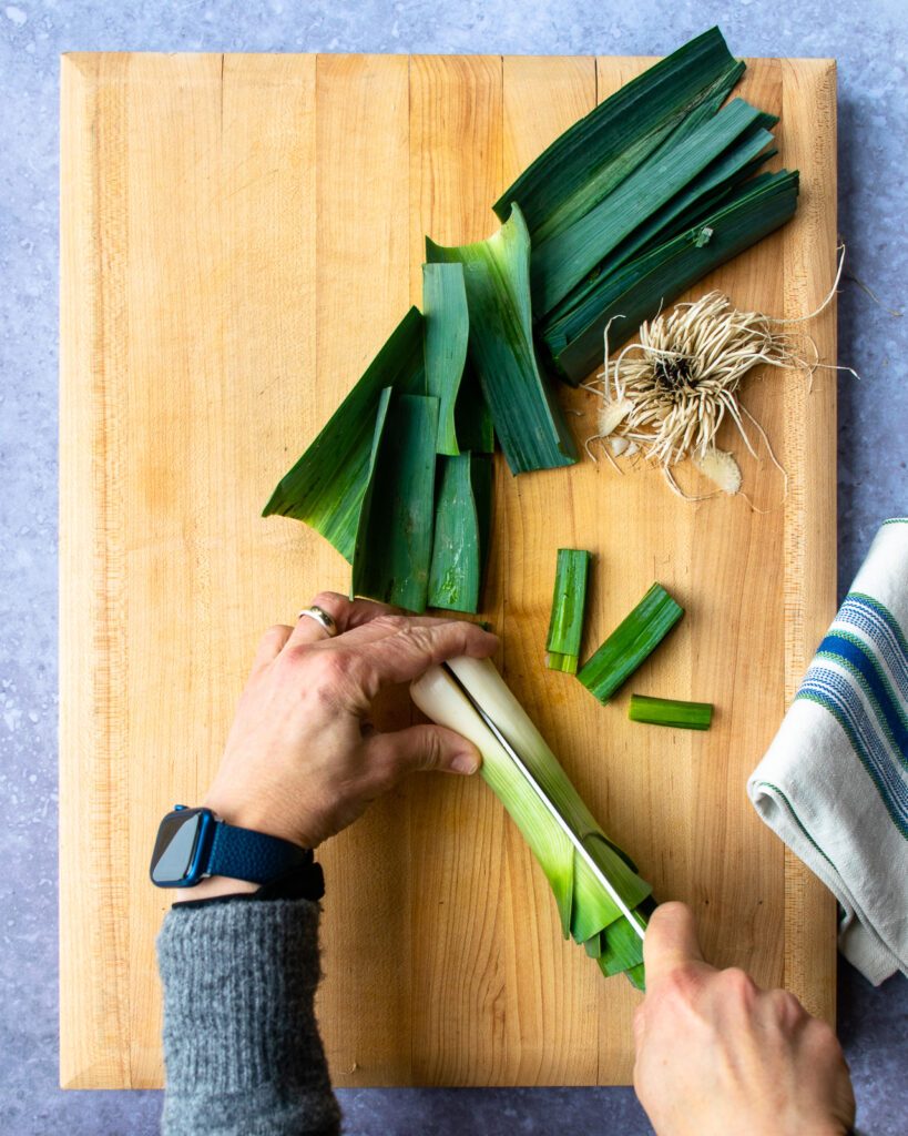 Hands slicing a leek in half lengthwise with a chef's knife.