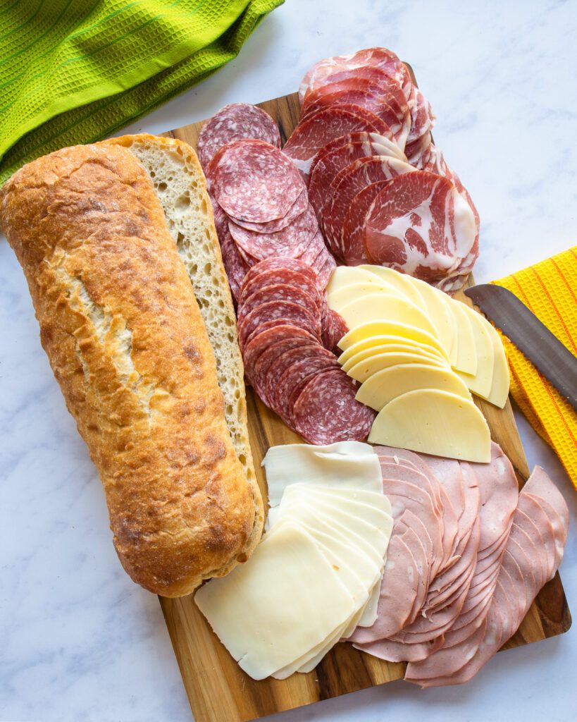 INgredients on a cutting board - a large sliced ciabatta loaf, cold cuts and sliced cheeses.