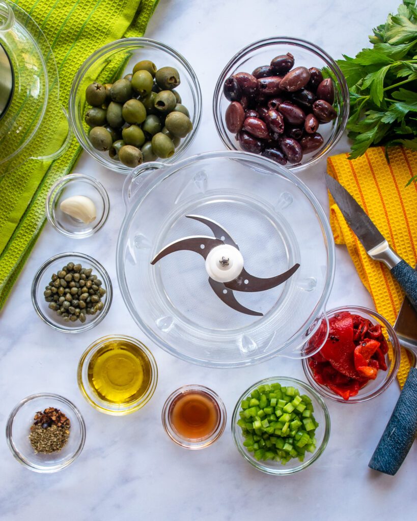 Ingredients surrounding a mini chopper - green and black olives, capers, garlic, celery, roasted red peppers, olive oil, vinegar, spices.