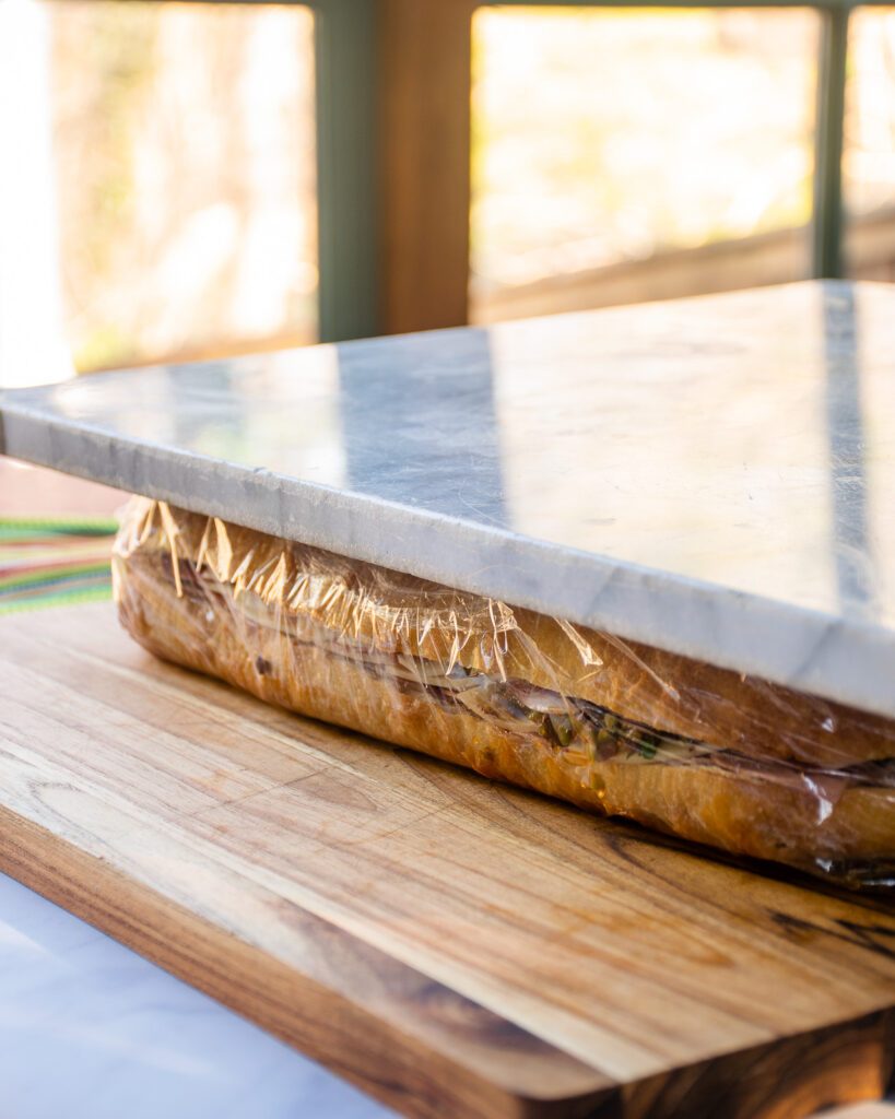 A marble slab pressing a large sandwich wrapped in plastic wrap.