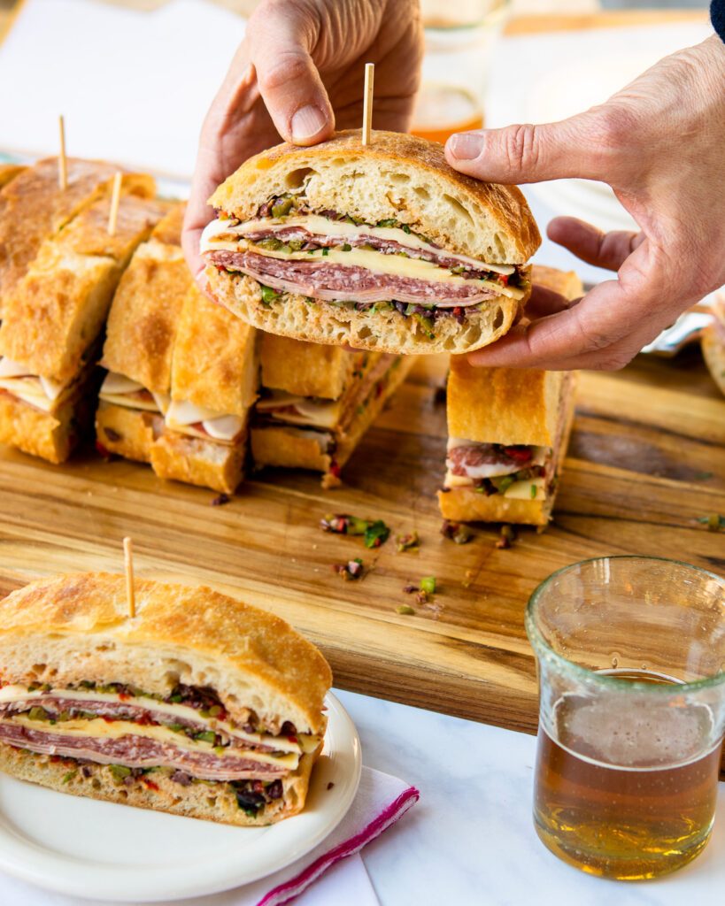 Hand holding a mini muffaletta sandwich over a cutting board of more sandwiches.