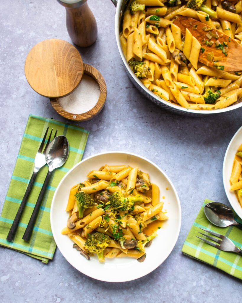 A white bowl with pasta, a green napkin, salt, pepper and more pasta in a sauteuse near by.