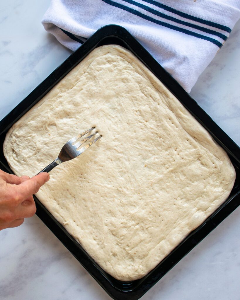 A fork docking pizza dough in a baking tray with a blue striped white kitchen towel near by.