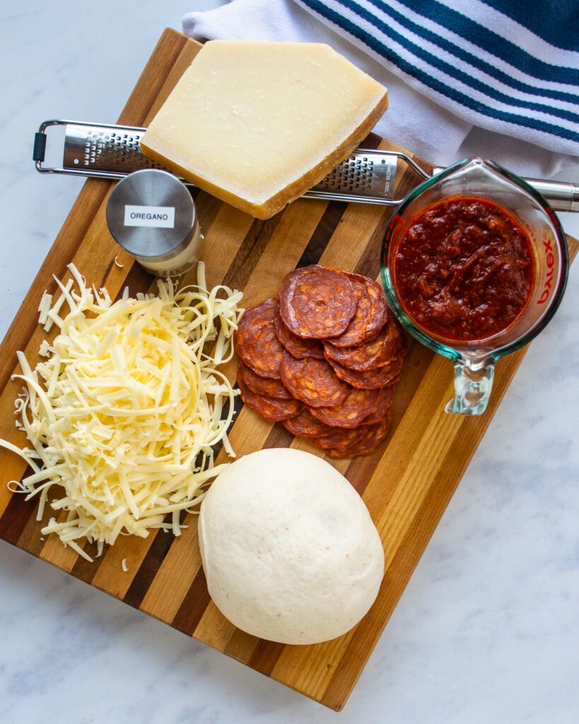 Ingredients for a pizza on a cutting board with a blue and white striped towel near by - oregano, grated cheese, pizza dough, pepperoni, pizza sauce and a block of Parmesan cheese.
