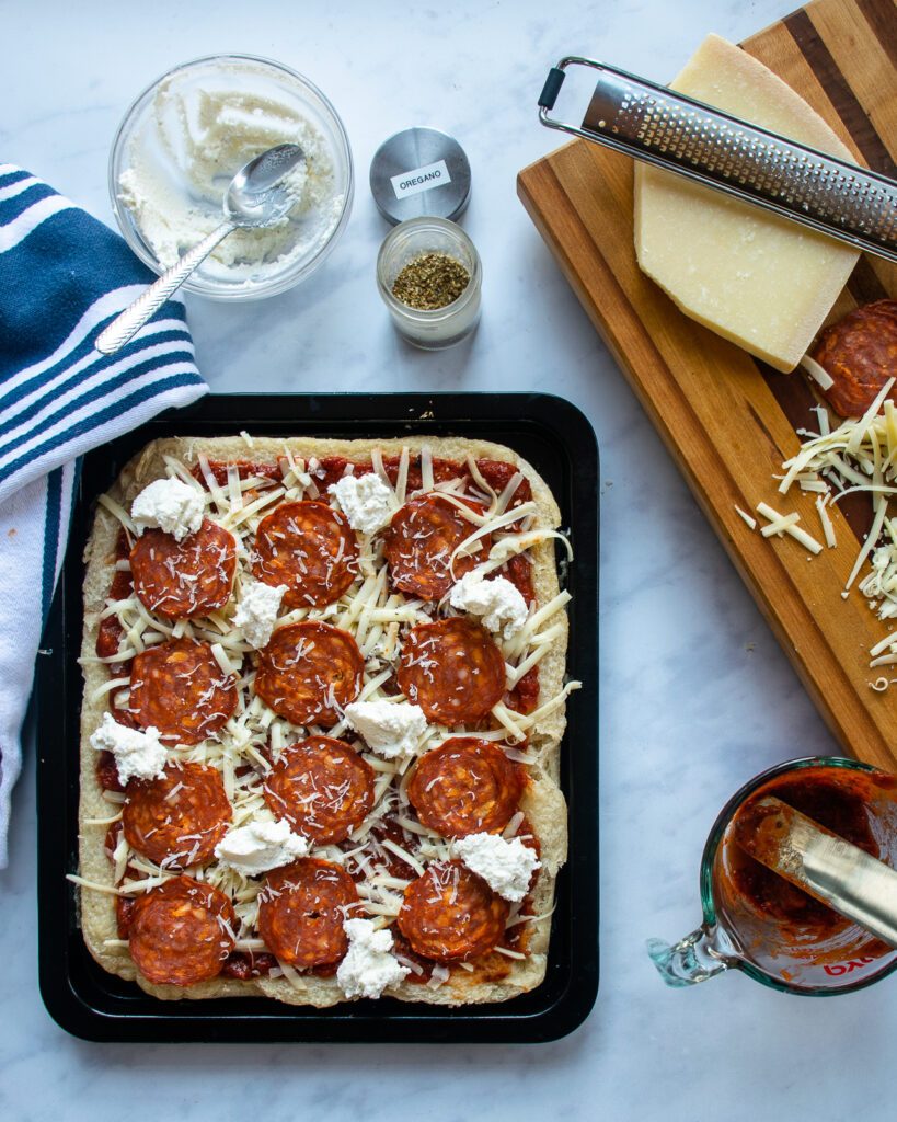 A raw pan pizza assembled on a baking sheet with pepperoni, ricotta, mozzarella, oregano and tomato sauce.