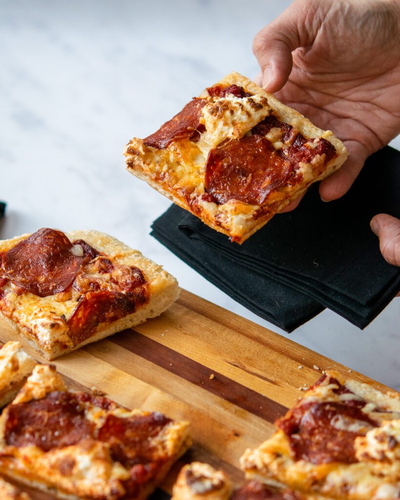 A hand holding up a piece of air fryer pan pizza above a cutting board with more pieces below.