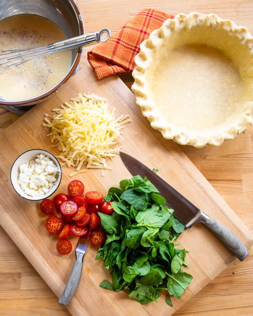An empty pie shell next to a cutting board with cheese, tomatoes, spinach and a bowl of egg custard.