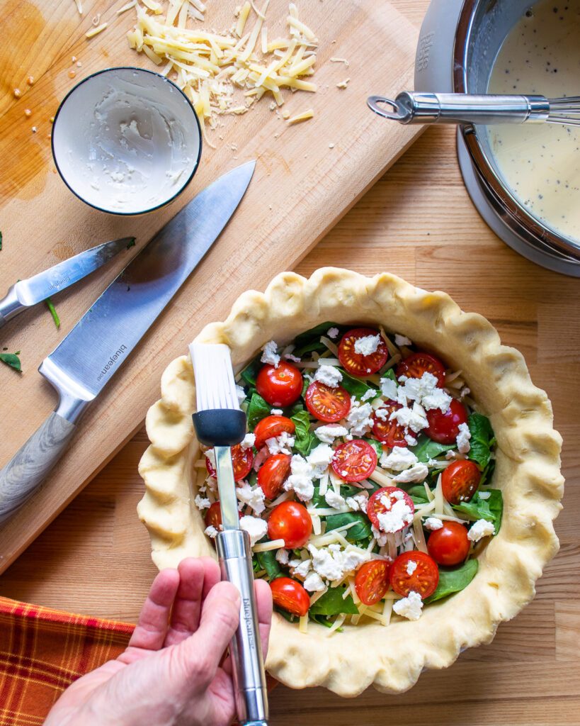 A hand brushing egg wash onto the rim of a pie crust with spinach, cheese and cherry tomatoes inside.