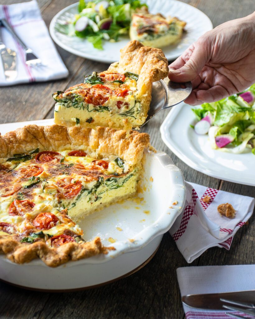 A hand lifting a wedge of Quiche Florentine out of a pie pan.