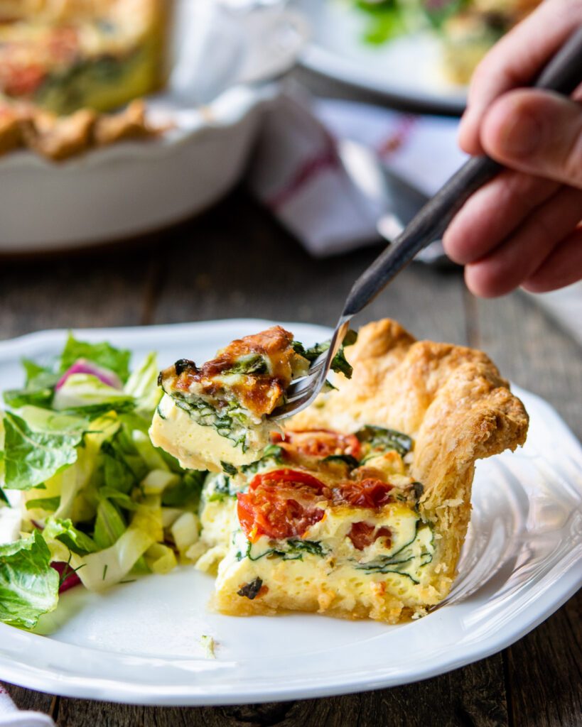 A hand forking a piece of quiche florentine out of a wedge on a white plate with a side salad.