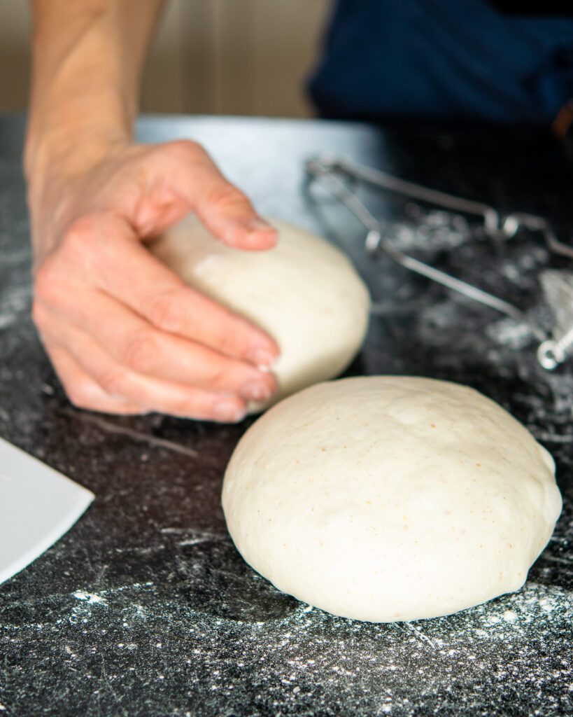A hand shaping dough on a countertop with a shaped ball of dough in the foreground.