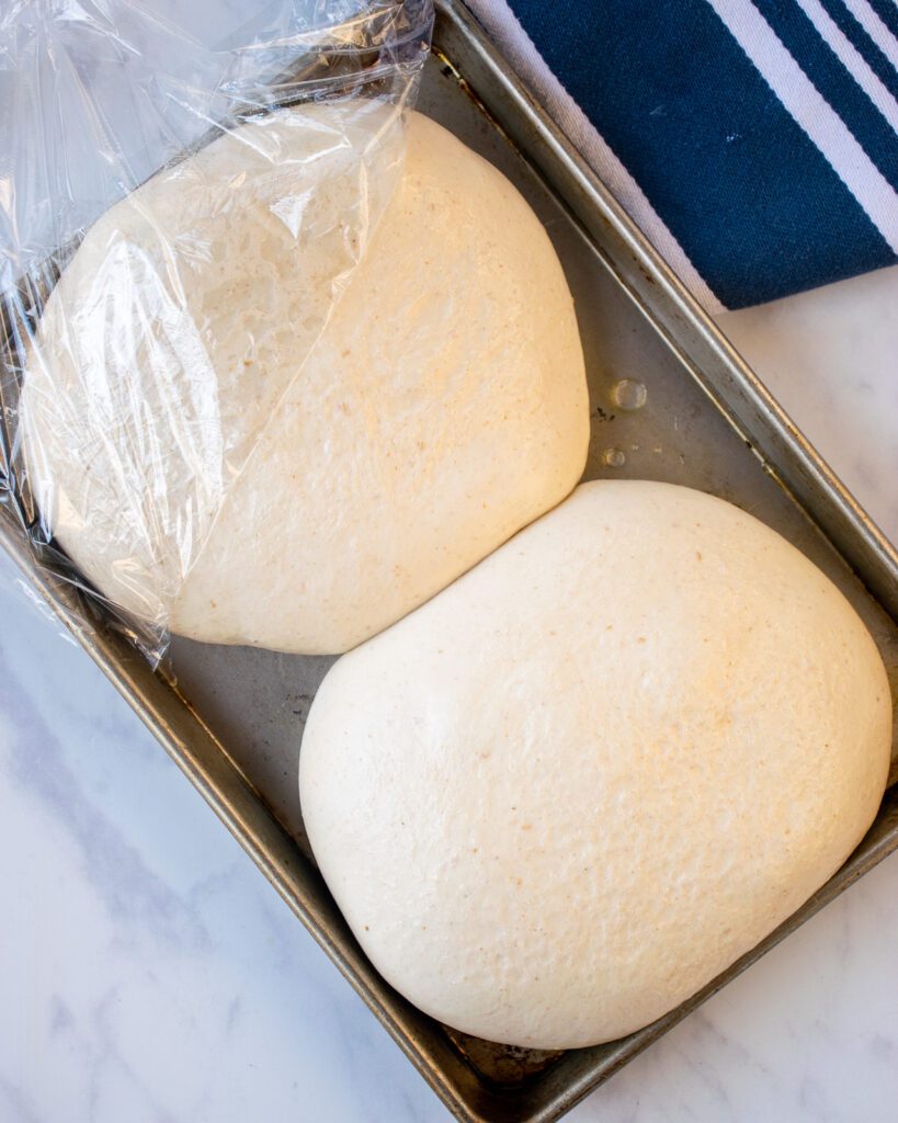 Two balls of risen dough on a baking sheet on a marble counter.