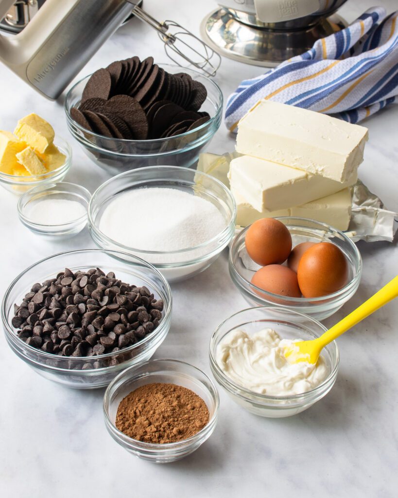 Ingredients on a marble countertop - sour cream, cocoa powder, chocolate chips, eggs, sugar, cream cheese, butter, chocolate wafers.
