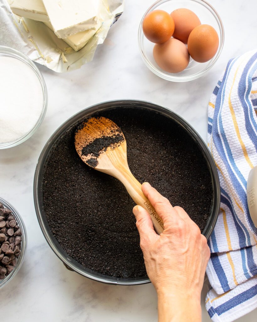 A hand pressing cookie crumbs down in a springform pan with a wooden spoon. A bowl of eggs, chocolate chips, cream cheese and a kitchen towel near by.