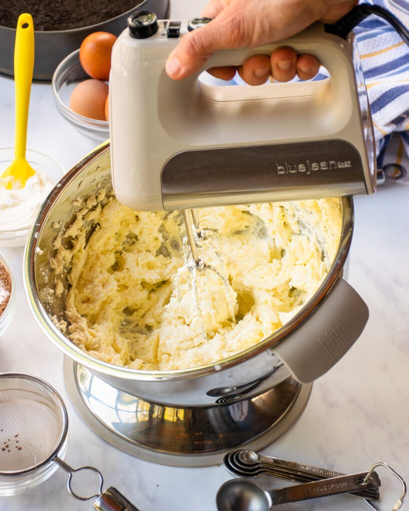 A hand mixer mixing cream cheese in a tilting stainless steel bowl with other ingredients around.