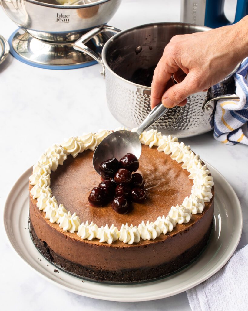 A hand spooning cherries onto the middle of a black forest cheesecake.