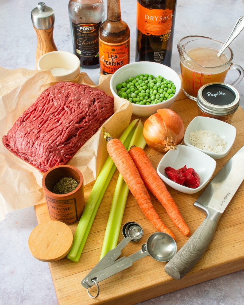 Ingredients on a cutting board - carrots, celery, onion, tomato paste, herbs, peas, ground beef.